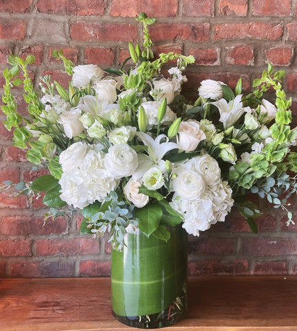 A large floral arrangement featuring white lilies, white roses, and green foliage in a tall green glass vase, set on a wooden surface against a red brick wall.