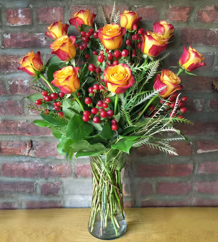 Bouquet of red and yellow roses with berries in a clear vase against a brick wall.
