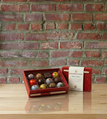 Red chocolate box with assorted chocolates and a card on a wooden surface against a brick wall.