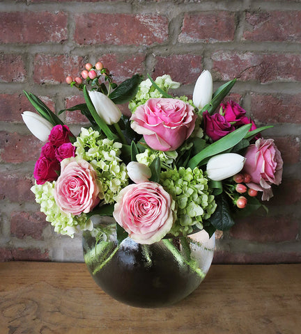 A floral arrangement featuring roses, spray roses, hydrangea, tulips, and berries in a glass vase, placed against a brick wall backdrop.