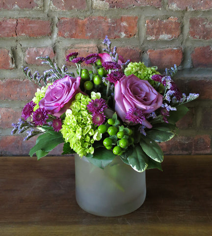 A floral arrangement featuring a cylinder vase with a mix of flowers including lavender roses, green hydrangea, hypericum berries, purple buttons, and purple filler, placed against a brick wall backdrop.